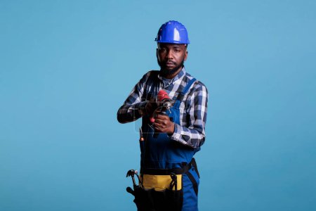 Photo for African american male holding a drill like a gun and pointing at the camera, serious attitude with construction tools on blue background. Builder wearing work uniform and tool belt. - Royalty Free Image