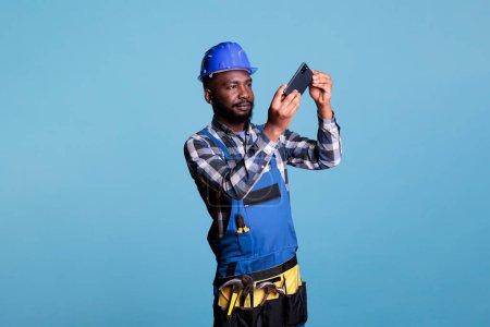 Téléchargez les photos : Travailleur de la construction afro-américain prenant des photos avec téléphone portable isolé sur fond bleu en studio tourné. Constructeur habillé en uniforme de travail distrait à l'aide d'un appareil mobile. - en image libre de droit