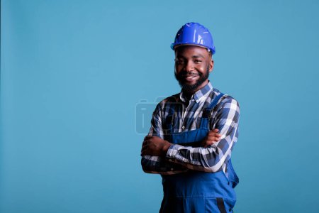 Téléchargez les photos : Constructeur afro-américain avec un visage heureux portant un casque souriant tout en regardant la caméra. Constructeur confiant posant avec les bras croisés sur fond bleu isolé. - en image libre de droit