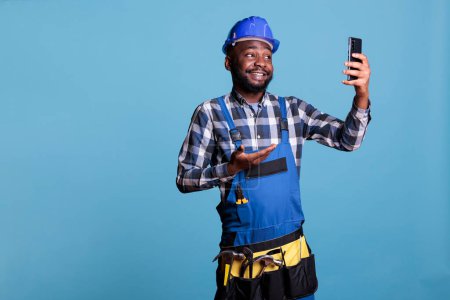 Téléchargez les photos : Entrepreneur afro-américain parlant sur téléphone portable appel vidéo sur fond bleu en studio tourné. Travailleur de la construction joyeux en salopette parler sur téléphone intelligent à propos d'un nouveau contrat de travail. - en image libre de droit