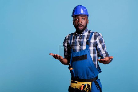 Photo for Portrait of confused african american builder posing in work uniform on blue background, making uncertain facial expression. Man shrugging shoulders, studio shot I dont know concept. - Royalty Free Image