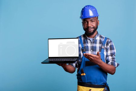 Téléchargez les photos : Portrait d'un joyeux constructeur afro-américain souriant à la caméra tout en tenant un ordinateur portable avec un écran vide debout dans un studio avec un fond bleu. Ouvrier de la construction en salopette - en image libre de droit