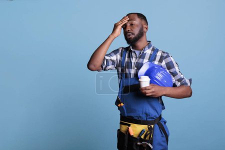 Photo for Exhausted construction employee drinking coffee during break. Overworked worker holding protective helmet wearing bibs while resting against blue background in studio shot. - Royalty Free Image