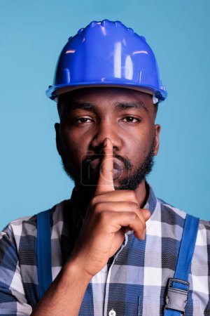 Photo for Portrait of male builder showing silence sign in front of camera, putting index finger over mouth to keep secret. Person making hush and mute gesture, confidential information in studio shot. - Royalty Free Image