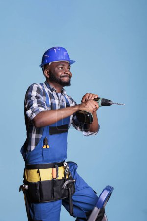 Téléchargez les photos : Entrepreneur afro-américain forant des clous avec perceuse électrique, travaillant avec une machine électrique sur le bâtiment de construction. Homme professionnel portant un hardhat avec des salopettes pour faire de la rénovation. - en image libre de droit