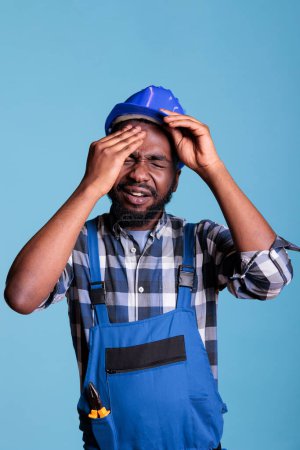 Photo for Construction worker suffering from a severe headache removing protective helmet. Contractor stressed by overwork touching his forehead with pained expression against blue background. - Royalty Free Image