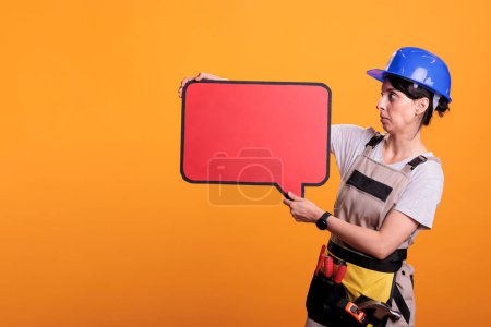 Photo for Female construction worker holding empty cardboard banner for advertisement, using carton copyspace. Woman builder showing red speech bubble with mockup on yellow background in studio. - Royalty Free Image