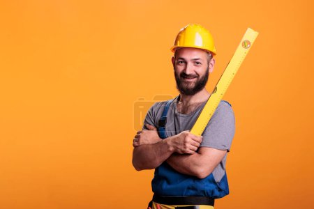 Photo for Repairing constructor holding water level ruler in studio, standing with arms crossed. Young repairman preparing to do measurements using professional leveler tool, confident carpenter. - Royalty Free Image