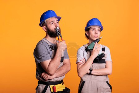 Photo for Team of builders brainstorming construction ideas in studio, thinking about renovating solution to start rebuilding work. Pensive man and woman holding power drilling gun and pair of pliers. - Royalty Free Image