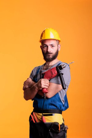 Photo for Professional builder posing on camera with power drill, acting confident before working on renovation. Handyman contractor using electric drilling nail gun over yellow background. - Royalty Free Image