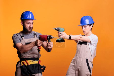 Photo for Team of construction workers holding drilling guns in studio, posing with electric screw tools over background. Man and woman using power drill and wearing overalls with hardhat, tools belt. - Royalty Free Image
