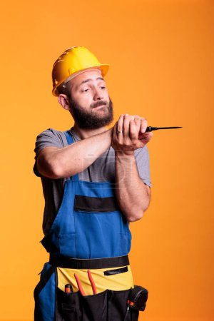 Photo for Construction worker using screwdriver to screw nails, carpenter dressed in overalls with hardhat and tools belt. Professional repairman using tool on yellow background in studio shot, industrial skill - Royalty Free Image