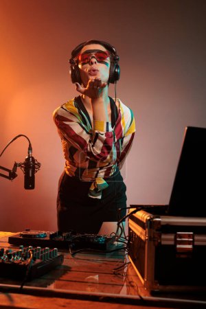 Téléchargez les photos : Femme DJ soufflant des baisers d'air en studio, étant romantique et agissant flirt alors qu'elle mélange de la musique techno aux platines tournantes. Charmant artiste rêveur essayant de remixer la chanson au mélangeur électrique. - en image libre de droit
