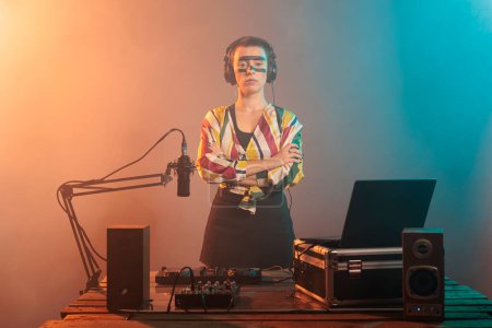 Photo for Female performer posing with arms crossed at turntables, using headphones and mixer to produce techno music or sounds. Doing performance with mixer and control buttons, audio stereo equipment. - Royalty Free Image