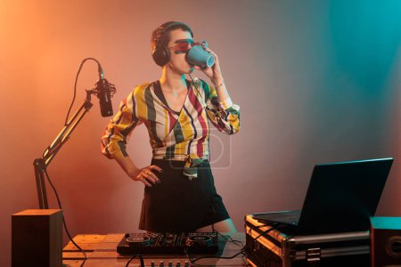 Photo for Cool DJ artist drinking coffee from mug before mixing techno music, preparing to do performance on turntables. Holding caffeine drink in cup and doing remix with volume mixer, energy beverage. - Royalty Free Image