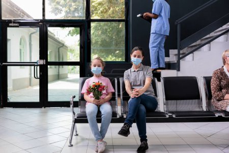 Photo for Child and mother wearing protective face mask to prevent infection with covid19 during medical appointment in hospital waiting area. Little girl holding flowers bouquet for grandmother - Royalty Free Image