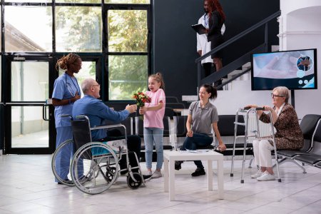 Photo for Granddaugher handing flowers bouquet to grandfather during checkup visit appointment in hospital waiting room. Disabled patient in wheelchiar discussing treatment with medical nurse. Medicine service - Royalty Free Image