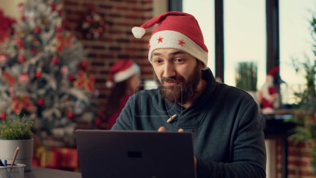 Photo for Male worker talking on videocall conference with webcam, wearing santa hat during christmas eve. Man chatting on online videoconference call in office with xmas holiday decorations. - Royalty Free Image