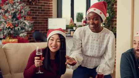 Foto de Grupo multiétnico de personas tintineando copas de vino en el evento de la fiesta de la oficina, sentado en el sofá y haciendo tostadas con alcohol para celebrar la víspera de Navidad. Ser festivo con bebida alcohólica el día de Navidad. - Imagen libre de derechos