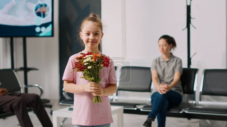 Téléchargez les photos : Jeune enfant souriant tenant un bouquet de fleurs dans la salle d'attente, debout dans le hall d'accueil de l'hôpital. Petite fille se sentant heureuse dans un centre de santé, attendant de rencontrer un médecin généraliste. - en image libre de droit