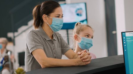 Photo for Mother and child with face mask talking to receptionist at hospital counter, people having medical appointment to see general practitioner. Woman and girl waiting at reception desk. Handheld shot. - Royalty Free Image