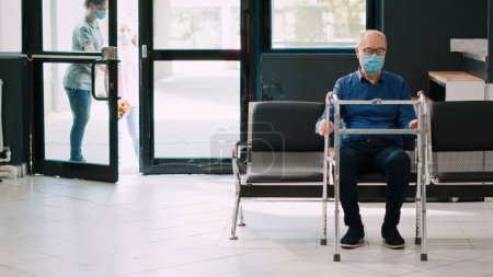 Foto de Madre y niña van a ver a un paciente anciano en silla de ruedas, visitando a una persona durante la pandemia covid 19. Personas con mascarilla en el vestíbulo del hospital para hablar con una anciana en la clínica. Disparo de mano. - Imagen libre de derechos