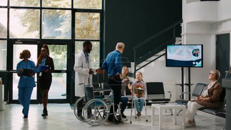 Foto de Hombre mayor en silla de ruedas tratando de caminar con el marco de caminar en el vestíbulo de la sala de espera, hablando con la mujer y el niño pequeño. Paciente anciano con discapacidad crónica que recibe tratamiento en el centro de salud. - Imagen libre de derechos