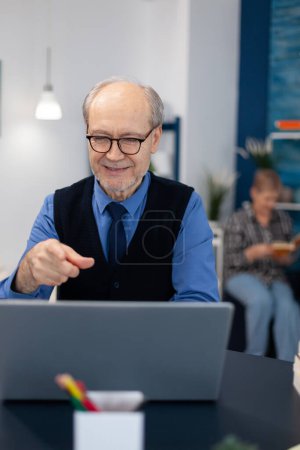 Téléchargez les photos : Heureux homme âgé pointant vers un ordinateur portable pendant la vidéoconférence. Pensionné joyeux avec les cheveux gris souriant à webcam ordinateur portable tout en bavardant assis au bureau . - en image libre de droit
