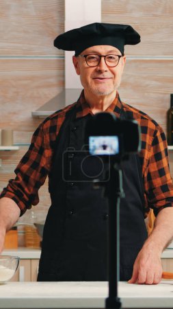 Photo for Experienced senior chef recording tutorial with food preparation in kitchen. Retired blogger influencer using internet technology communicating shooting blogging on social media with digital equipment - Royalty Free Image