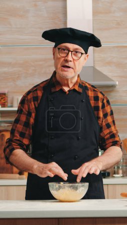 Photo for Old man wearing bonete while explaining food preparation recipe looking at camera. Retired blogger chef influencer using internet technology communicating on social media with digital equipment - Royalty Free Image