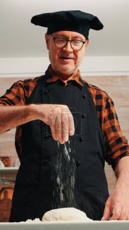 Photo for Senior man adding flour on dough by hand looking at camera smiling. Retired elderly chef with bonete and uniform sprinkling, sieving spreading rew ingredients with hand baking homemade pizza and bread - Royalty Free Image