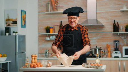 Photo for Old man with kitchen apron playing with bread dough at home smiling in front of camera. Retired elderly chef forming pizza countertop on a floured surface and kneading it with hands, in modern kitchen - Royalty Free Image