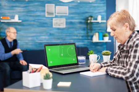 Téléchargez les photos : Femme âgée confiante utilisant un ordinateur portable Femme âgée travaillant sur un ordinateur avec écran vert et mari tenant télécommande tv. - en image libre de droit