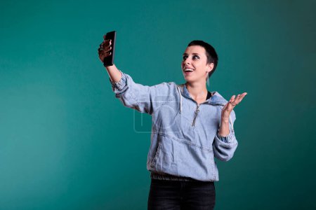 Téléchargez les photos : Femme caucasienne joyeuse souriant tout en parlant avec un ami distant lors d'une réunion de vidéocall en ligne au téléphone, debout dans un studio avec un arrière-plan isolé. Femme bavarder avec l'équipe sur vidéoconférence - en image libre de droit