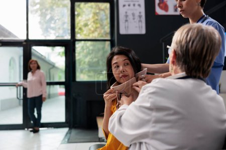 Téléchargez les photos : Traumatologue médecin et infirmière aidant le patient âgé à enlever le collier cervical lors d'un rendez-vous d'examen dans la salle d'attente de l'hôpital. Femme asiatique recevant un soutien médical - en image libre de droit