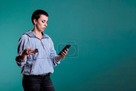 Photo for Portrait of confident woman using credit card to do online shopping using phone in studio, making electronic payment transaction on retail store website. Cheerful female buying on sale discount with - Royalty Free Image