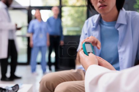 Photo for Senior caucasian medic measuring sugar level with blood sample to do insulin test with glucometer for asian patient with diabetes during consultation. Doctor checking glucose in hospital waiting area - Royalty Free Image