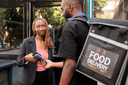 Photo for Office worker paying for food delivery with smartphone, pos terminal contactless payment, nfc technology. African american courier delivering restaurant meal to cheerful customer - Royalty Free Image