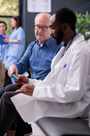 Téléchargez les photos : Médecin afro-américain consultant un patient âgé avec un instrument de glucomètre tout en mesurant le taux d'insuline et de glucose à partir d'un échantillon de sang. Homme âgé ayant une consultation de visite de contrôle dans le lobby de l'hôpital - en image libre de droit
