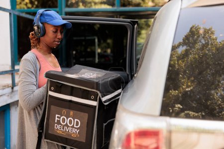 Téléchargez les photos : Courrier afro-américain dans les écouteurs livrant de la nourriture à emporter en voiture, en attendant le client près de l'immeuble de bureaux à l'extérieur. Travailleur de service de livraison sortant sac à dos de l'automobile - en image libre de droit