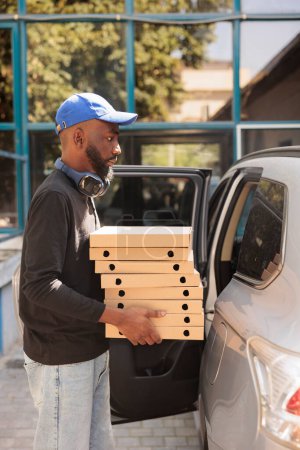 Téléchargez les photos : Coursier afro-américain transportant pizza au bureau en voiture vue de côté, livreur tenant des boîtes pile. Déjeuner livreur de services portant fastfood, debout près du bâtiment de l'entreprise à l'extérieur - en image libre de droit
