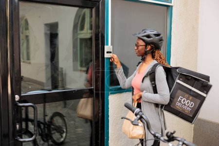 Téléchargez les photos : Femme livrant de la nourriture, pressant la sonnette de l'immeuble de bureaux, attendant le client à l'extérieur. Restaurant afro-américain à emporter service de livraison de repas courrier debout devant la porte - en image libre de droit