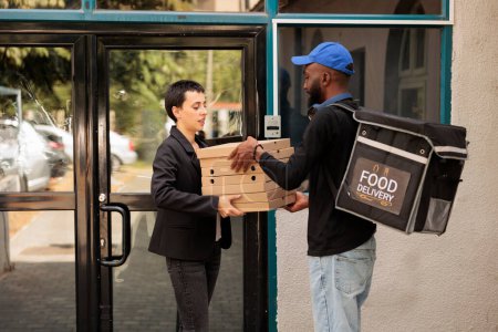 Téléchargez les photos : Courrier afro-américain donnant commande de pizza client à l'extérieur, femme prenant des paquets de repas pile pour l'équipe de travailleurs de bureau. Jeune homme avec sac à dos thermique offrant un déjeuner à emporter - en image libre de droit