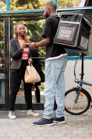 Photo for Office worker paying for food delivery with credit card, pos terminal contactless payment for order. African american courier with backpack delivering restaurant meal to customer outdoors - Royalty Free Image