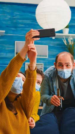 Photo for Cheerful happy mixed race friends taking selfie wearing face mask keeping social distancing against spread of virus sitting on couch in living room. Diverse people socializing keeping social distance - Royalty Free Image