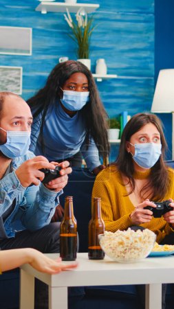 Téléchargez les photos : Des amis multiraciaux concentrés jouant à des jeux vidéo avec manette dans le salon portant un masque facial pour empêcher la propagation du coronavirus. Diverses personnes s'amusent, riant à la nouvelle fête normale - en image libre de droit