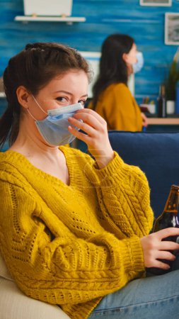 Photo for Woman holding beer bottle drinking looking at camera, relaxing on sofa, spending time with friends keeping social distancing with face mask in living room preventing spread of coronavirus. - Royalty Free Image