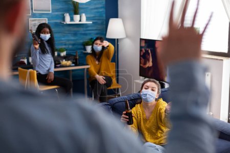 Photo for Man coming at party with friends wearing face mask preventing spread of coronavirus, women holding beer bottles socializing in apartment living room. People enjoying time during covid 19 outbreak - Royalty Free Image