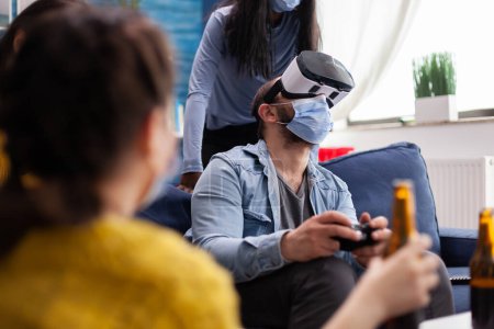 Photo for Excited man spending time with friends experiencing virtual reality playing games with vr headset wearing face mask to prevent coronavirus spread in global pandemic. Diverse people having fun at new - Royalty Free Image