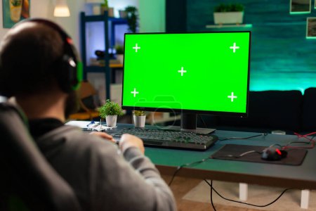Téléchargez les photos : Cyberjoueur professionnel jouant à des jeux vidéo numériques sur un ordinateur puissant avec écran vert. Joueur utilisant pc avec chroma mock up isolé jeux de tir de streaming de bureau portant casque - en image libre de droit